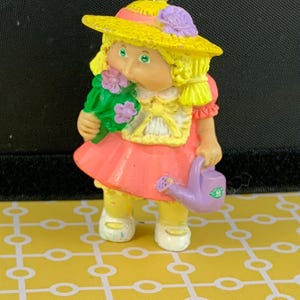 May include: A vintage figurine of a girl in a yellow hat with purple details and a pink dress. She holds a bouquet and a purple watering can. The figurine has blonde hair and wears yellow shoes. A collectible item.
