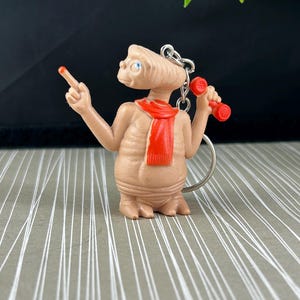 May include: A tan-coloured E.T. figurine keyring with a red scarf and a red finger. The keyring is holding a red device in one hand and pointing with the other. The background is a grey and white striped pattern.