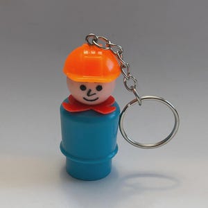 May include: A vintage Fisher-Price Little People keychain featuring a construction worker figure. The figure has a blue body, orange hard hat, and a red collar. The keychain includes a silver chain and a round key ring.