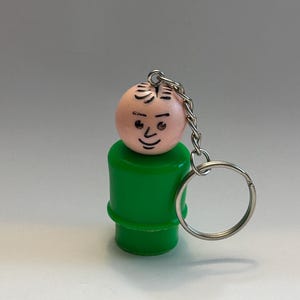 May include: A vintage-style keychain featuring a classic Little People figure. The figure has a light pink head with a painted face and a green body. The keychain includes a silver-colored chain and a split ring.