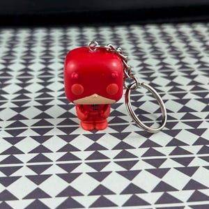 May include: A red vinyl keychain figure with a silver chain and keyring. The figure has a square head, small eyes, and a white chin. The figure is wearing a red outfit. The background is a white surface with a black triangle pattern.