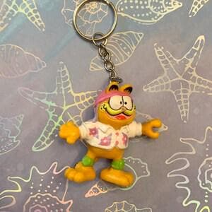 Peut inclure: Un porte-clés Garfield représentant le chat de dessin animé portant une chemise hawaïenne, un chapeau rose et un pantalon vert. La figurine jaune a les bras tendus et une expression souriante. Le porte-clés est attaché à un anneau et une chaîne argentés.