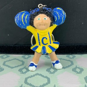 Op de afbeelding: Een Cabbage Patch Kids cheerleader sleutelhanger. Het figuur draagt een geel en blauw uniform met de letter "C" op de voorkant. De cheerleader heeft zwart haar, groene ogen en houdt blauwe pompons vast. De sleutelhanger is van plastic.