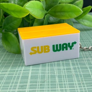 以下が含まれることがあります： 白と黄色のSubwayブランドのキーホルダー。キーホルダーには、緑と黄色のSubwayのロゴが描かれています。背景には緑の葉と、薄い緑色の模様のある表面が含まれています。