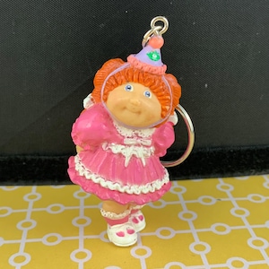 Peut inclure: Un porte-clés représentant une poupée Cabbage Patch Kids. La poupée porte une robe rose avec des bordures blanches, des chaussures blanches et un chapeau de fête violet. La poupée a des cheveux roux et des yeux bleus. Le porte-clés est attaché à un anneau argenté.