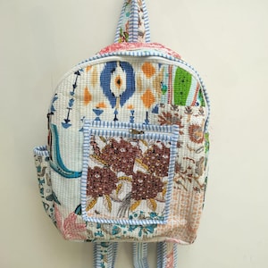Op de afbeelding: Een gewatteerde rugzak met een patchworkontwerp met diverse patronen, waaronder blauwe en oranje ikat, groene botanische prints en een voorvak met een bruin bloemmotief. De tas is voorzien van lichtblauw en wit gestreepte biezen en schouderbanden.