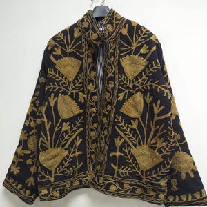 May include: A black jacket featuring intricate gold-colored floral embroidery throughout the front and sleeves. The garment has a high collar, long sleeves, and a visible black and white striped lining visible at the neck opening.