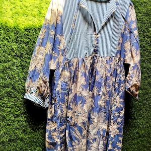 Può includere: Abito a maniche lunghe con corpetto a righe blu e bianche e gonna con stampa floreale blu. L'abito è appeso a una gruccia di legno. Le maniche hanno un motivo a righe.