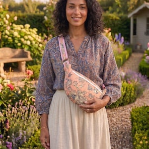 May include: A woman in a paisley blouse and cream skirt, holding a peach-coloured fanny pack with a floral pattern. The fanny pack has a pink and white striped strap. The background shows a garden with flowers and a small shed.