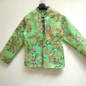 Puede incluir: Chaqueta acolchada verde claro con un estampado floral en tonos rosa, amarillo y verde. La chaqueta tiene dos bolsillos delanteros y un cuello mandarín. La chaqueta está colgada de una percha negra.