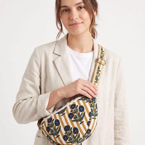 May include: A crescent-shaped crossbody bag featuring a mustard yellow and white vertical striped pattern with dark blue floral motifs. The bag includes an adjustable matching striped fabric strap with gold-toned metal hardware and a zip closure.