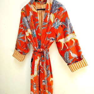 May include: An orange robe with a jungle print featuring leopards, zebras, and tropical foliage. The robe has a striped interior and cuffs, and a matching belt. The robe is hanging on a black hanger.