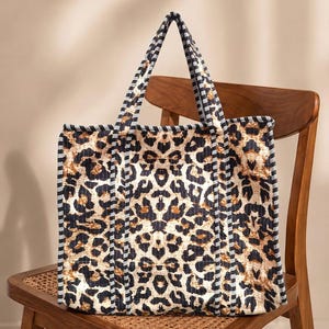 May include: A square-shaped tote bag featuring a bold black and tan leopard print pattern. The bag is constructed with a quilted texture and is finished with black and white striped fabric trim along the edges and the two top carry handles.