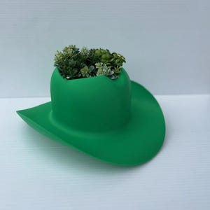 May include: A vibrant green cowboy hat planter filled with small, green and white artificial plants. The hat is a solid, bright green color and sits on a white surface. The hat's brim is wide and curved, and the crown holds the greenery.