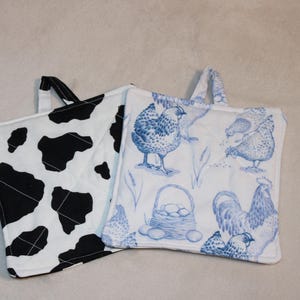 May include: Two square pot holders. One features a black and white cow print. The other has a blue and white chicken design with a basket of eggs. Both have a small fabric loop for hanging.
