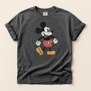 K&ouml;nnte beinhalten: Dunkelgraues T-Shirt mit einem Vintage-Motiv von Micky Maus. Die Zeichentrickfigur ist in ihrer klassischen Kleidung dargestellt: rote Shorts, gelbe Schuhe, wei&szlig;e Handschuhe und schwarze Ohren. Das T-Shirt hat hochgekrempelte &Auml;rmel und ein Comfort Colors-Etikett.