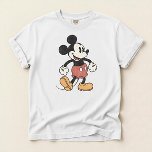 K&ouml;nnte beinhalten: Wei&szlig;es T-Shirt mit einer Vintage-Illustration von Mickey Mouse. Die Zeichentrickfigur ist in Schwarz, Rot und Gelb dargestellt, mit einem Used-Effekt. Das T-Shirt hat hochgekrempelte &Auml;rmel und ein Comfort Colors-Etikett.