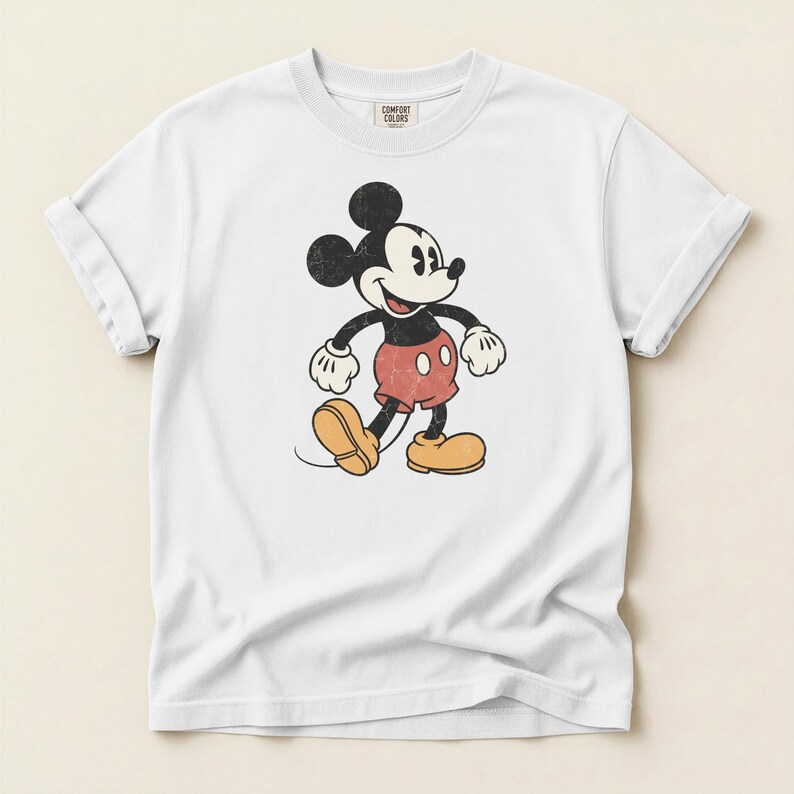 K&ouml;nnte beinhalten: Wei&szlig;es T-Shirt mit einer Vintage-Illustration von Mickey Mouse. Die Zeichentrickfigur ist in Schwarz, Rot und Gelb dargestellt, mit einem Used-Effekt. Das T-Shirt hat hochgekrempelte &Auml;rmel und ein Comfort Colors-Etikett.
