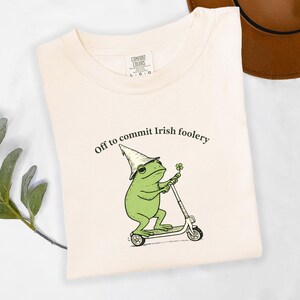 May include: Cream-colored t-shirt with the text "Off to commit Irish foolery." The shirt features a green frog wearing a leprechaun hat and riding a scooter, holding a four-leaf clover. The shirt is a Comfort Colors brand.