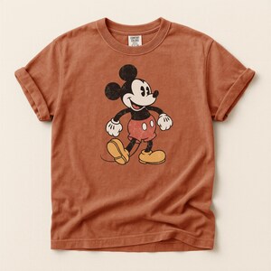 K&ouml;nnte beinhalten: Ein rostfarbenes T-Shirt mit einer Vintage-Grafik von Mickey Mouse. Die Zeichentrickfigur ist in Schwarz, Wei&szlig;, Rot und Gelb dargestellt. Das Shirt hat hochgekrempelte &Auml;rmel und einen Rundhalsausschnitt. Das Etikett lautet "Comfort Colors".