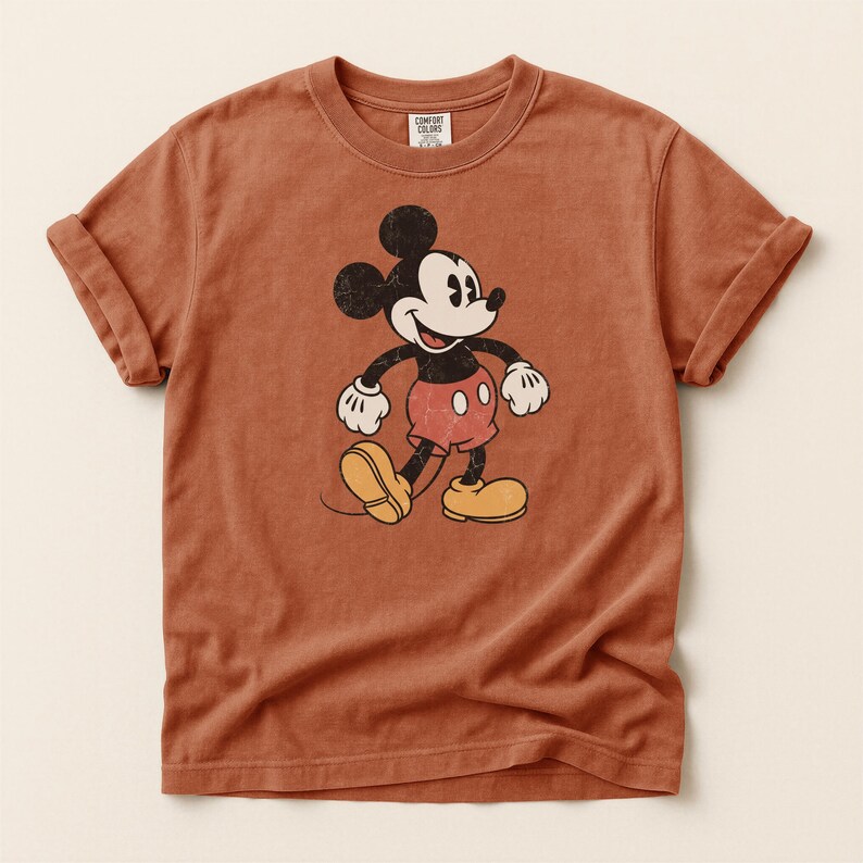 K&ouml;nnte beinhalten: Ein rostfarbenes T-Shirt mit einer Vintage-Grafik von Mickey Mouse. Die Zeichentrickfigur ist in Schwarz, Wei&szlig;, Rot und Gelb dargestellt. Das Shirt hat hochgekrempelte &Auml;rmel und einen Rundhalsausschnitt. Das Etikett lautet "Comfort Colors".