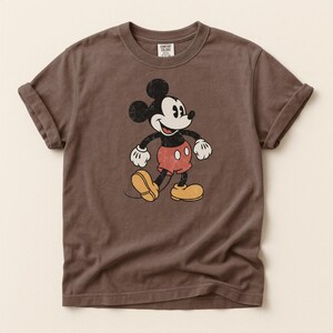 K&ouml;nnte beinhalten: Braunes T-Shirt mit einem verblichenen Mickey Mouse-Motiv. Die Zeichentrickfigur ist in Schwarz, Wei&szlig;, Rot und Gelb dargestellt. Das Shirt hat hochgekrempelte &Auml;rmel und einen Rundhalsausschnitt. Das Etikett lautet "Comfort Colors".