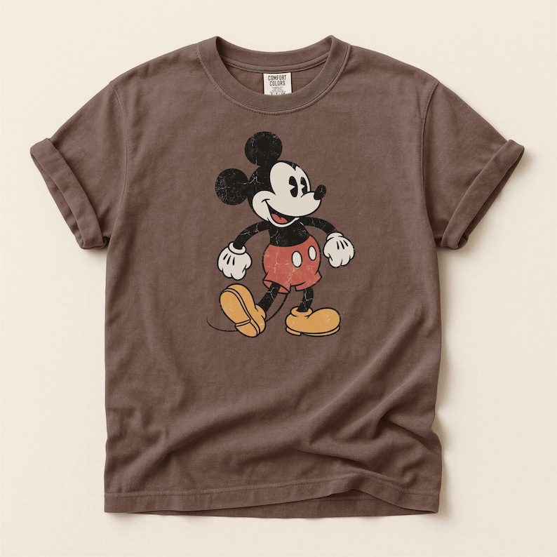 K&ouml;nnte beinhalten: Braunes T-Shirt mit einem verblichenen Mickey Mouse-Motiv. Die Zeichentrickfigur ist in Schwarz, Wei&szlig;, Rot und Gelb dargestellt. Das Shirt hat hochgekrempelte &Auml;rmel und einen Rundhalsausschnitt. Das Etikett lautet "Comfort Colors".