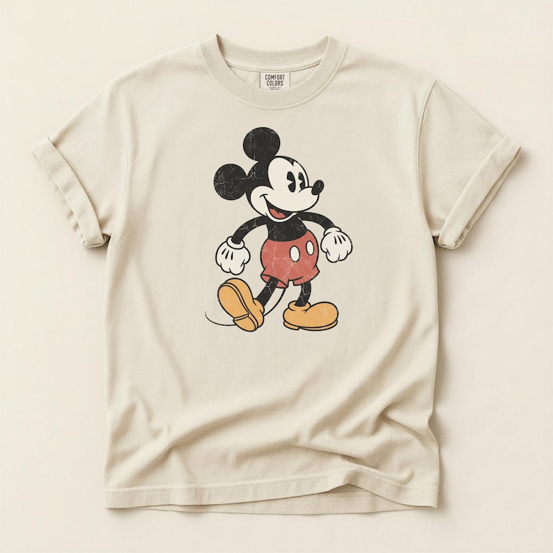 K&ouml;nnte beinhalten: Cremefarbenes T-Shirt mit einem Vintage-Mickey-Mouse-Motiv. Die Zeichentrickfigur ist in Schwarz, Rot und Gelb dargestellt, mit einem Used-Effekt. Das Shirt hat hochgekrempelte &Auml;rmel und der Text "COMFORT COLORS" ist oben links aufgedruckt.