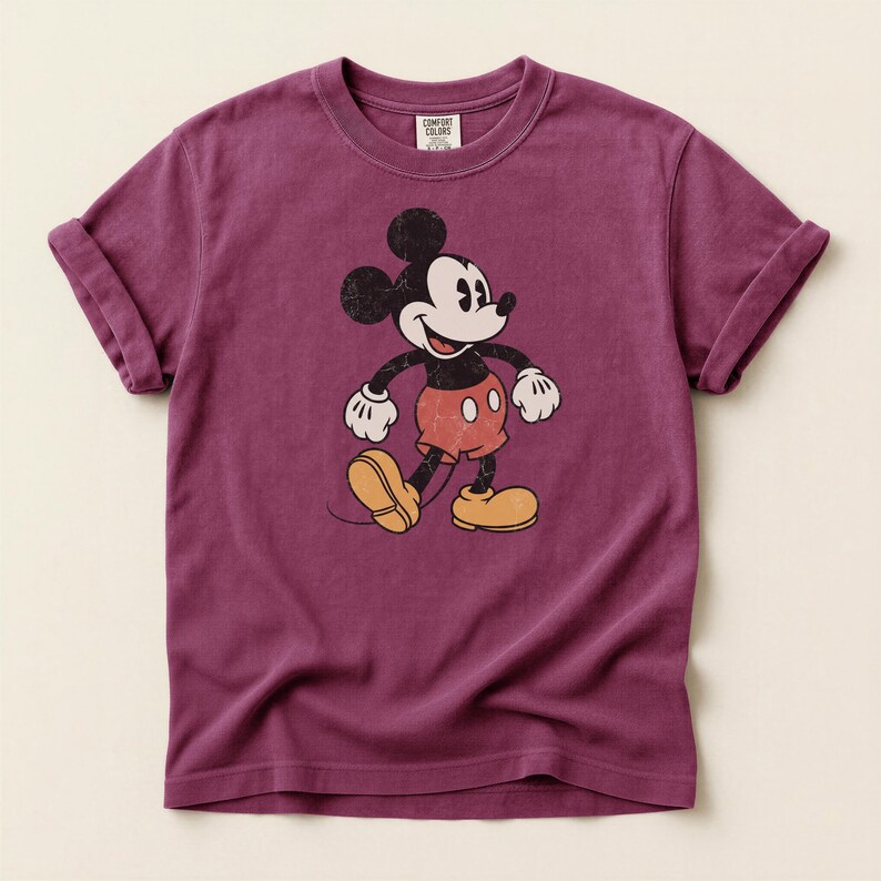 K&ouml;nnte beinhalten: Ein weinrotes T-Shirt mit einem Vintage-Grafik von Mickey Mouse. Die Zeichentrickfigur ist in ihrem klassischen Outfit mit roten Shorts, gelben Schuhen und wei&szlig;en Handschuhen dargestellt. Das T-Shirt hat hochgekrempelte &Auml;rmel und das Comfort Colors-Logo.