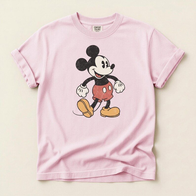 K&ouml;nnte beinhalten: Hellrosa T-Shirt mit einem Vintage-Grafik von Mickey Mouse. Die Zeichentrickfigur ist in ihrem klassischen Outfit mit roten Shorts, gelben Schuhen und wei&szlig;en Handschuhen dargestellt. Das Shirt hat hochgekrempelte &Auml;rmel und den Text "COMFORT COLORS".