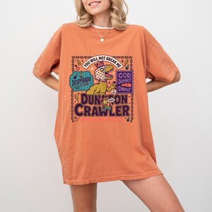 May include: An oversized burnt orange t-shirt featuring a graphic print of a dinosaur wearing a heart-patterned headscarf and crown. The text reads "YOU WILL NOT BREAK ME", "DESPERADO CLUB", "GOD DAMNIT DONUT", and "DUNGEON CRAWLER" in bold lettering.