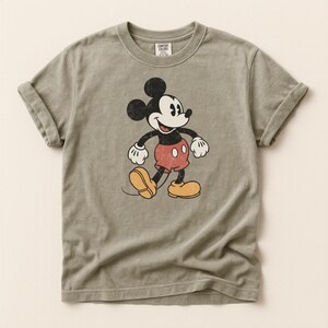 K&ouml;nnte beinhalten: Salbeigr&uuml;nes T-Shirt mit einem verblichenen Mickey Mouse-Motiv. Die Zeichentrickfigur ist in Schwarz, Wei&szlig;, Rot und Gelb dargestellt. Das Shirt hat hochgekrempelte &Auml;rmel und einen Rundhalsausschnitt. Das Etikett lautet "Comfort Colors".