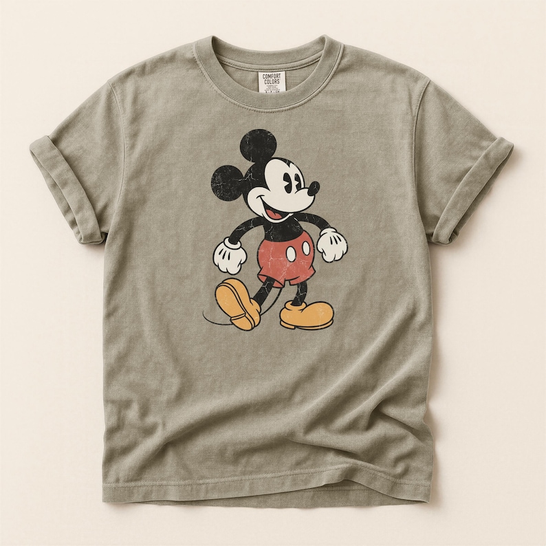 K&ouml;nnte beinhalten: Salbeigr&uuml;nes T-Shirt mit einem verblichenen Mickey Mouse-Motiv. Die Zeichentrickfigur ist in Schwarz, Wei&szlig;, Rot und Gelb dargestellt. Das Shirt hat hochgekrempelte &Auml;rmel und einen Rundhalsausschnitt. Das Etikett lautet "Comfort Colors".