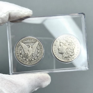 May include: Two silver Morgan dollar coins, one dated 1889, displayed in a clear plastic holder. The coins feature detailed engravings of an eagle and a woman's profile. The holder is held by a gloved hand.