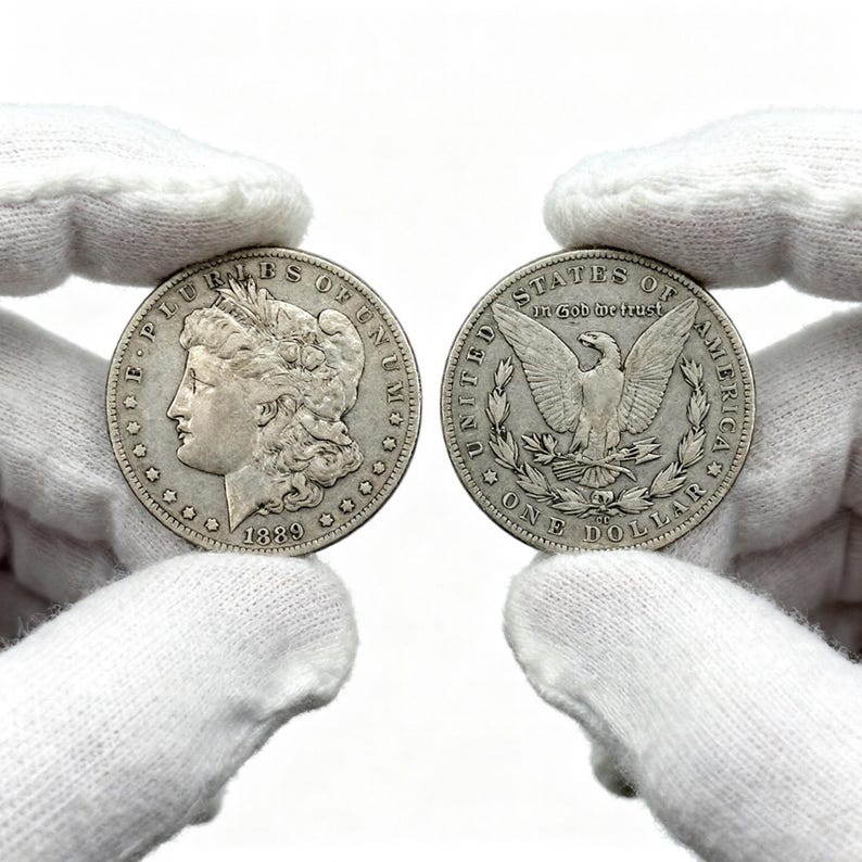 May include: Two silver Morgan silver dollars, one dated 1889, are displayed. The obverse of the coin features a profile of a woman and the words "E PLURIBUS UNUM". The reverse shows an eagle with the words "UNITED STATES OF AMERICA" and "ONE DOLLAR".