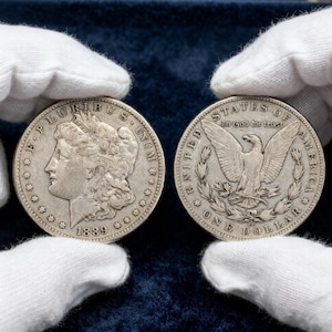 Puede incluir: Se muestran dos monedas de plata Morgan, una de 1889. El anverso presenta el perfil de una mujer y la inscripción "E PLURIBUS UNUM". El reverso muestra un águila con las palabras "UNITED STATES OF AMERICA" y "ONE DOLLAR".
