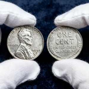 以下が含まれることがあります： 1944年製の銀色の米国の1セント硬貨2枚が、手袋をはめた手で持たれています。1枚の硬貨にはアブラハム・リンカーンの横顔と「LIBERTY」の文字が描かれています。もう1枚の硬貨には「ONE CENT」と「UNITED STATES OF AMERICA」と書かれています。