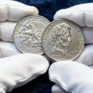Puede incluir: Se muestran dos monedas de plata, una con un águila y las palabras "Half Dollar", la otra con un retrato y el año 1918. Las monedas son sostenidas por manos enguantadas sobre un fondo azul oscuro.