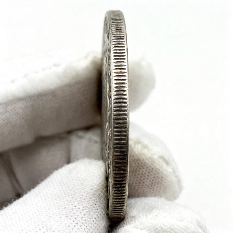 May include: A silver coin, viewed from the side, showcasing a detailed, ridged edge. The coin's surface displays intricate designs and text. The coin is held by a gloved hand against a white background, highlighting its metallic sheen.