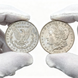 Puede incluir: Dos dólares de plata Morgan, uno con un águila con el texto "In God We Trust" y "One Dollar", el otro con un perfil de mujer y el año 1881. Las monedas son sostenidas por manos enguantadas.
