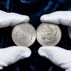 Op de afbeelding: Twee zilveren Morgan dollar munten, een met een adelaar en de woorden "United States of America" en "One Dollar". De andere munt toont een vrouwenprofiel en de datum 1878. Beide munten worden vastgehouden door gehandschoende handen.