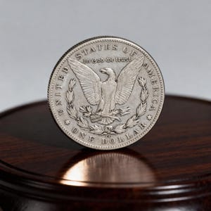 May include: A silver One Dollar coin with an eagle design, the words "In God We Trust" and "United States of America" are visible. The coin is on a dark wooden surface.