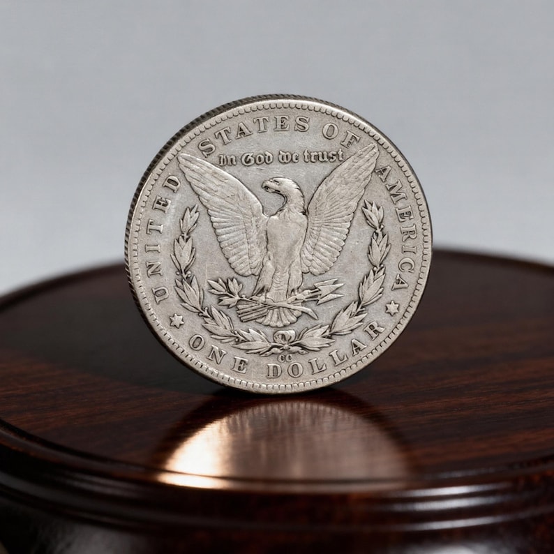 May include: A silver One Dollar coin with an eagle design, the words "In God We Trust" and "United States of America" are visible. The coin is on a dark wooden surface.