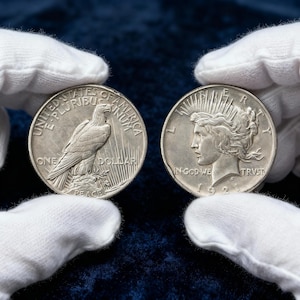 May include: Two silver Peace Dollars, each held by a gloved hand. One coin shows an eagle and the words "ONE DOLLAR" and "PEACE". The other coin features a woman's profile and the words "IN GOD WE TRUST" and the date "1921".