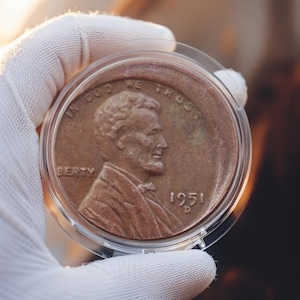 Puede incluir: Un centavo de cobre de 1951 dentro de un soporte de plástico transparente. La moneda muestra el perfil de Abraham Lincoln y la inscripción "IN GOD WE TRUST". El año "1951" es visible.