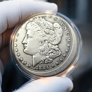 Può includere: Una moneta d'argento Morgan del 1884, racchiusa in una capsula protettiva circolare in plastica trasparente. Il dritto presenta il profilo di Lady Liberty rivolto a sinistra, con il testo LIBERTY, E PLURIBUS UNUM e l'anno 1884 visibili.