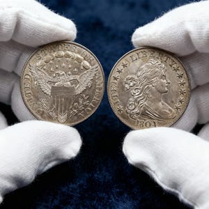 May include: Two antique silver coins are held by gloved hands. One coin features an eagle with outstretched wings, the other depicts a woman's profile with the year 1804. The coins are detailed with stars and text.