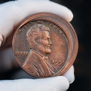 May include: A large, copper-colored coin featuring a profile of a man, with the text "IN GOD WE TRUST" and the year "1955". The coin is held by a gloved hand.
