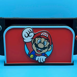 May include: A black Nintendo Switch console sits in a red and black stand featuring a cartoon image of Mario. The stand has a light blue border and is set against a blue background. The console's controllers are visible on either side.