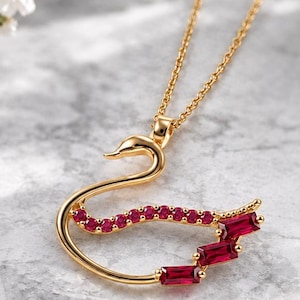 May include: A gold-toned necklace featuring a swan-shaped pendant. The swan's body is adorned with a curved line of small round red stones and three rectangular red baguette-cut stones forming the wing, suspended from a delicate gold chain.